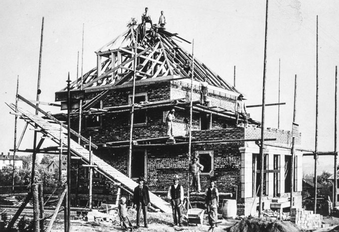 Cantiere con un ponteggio di travi tonde, assi e corde. Il materiale viene trasportato sui vari livelli del ponteggio tramite una rampa.